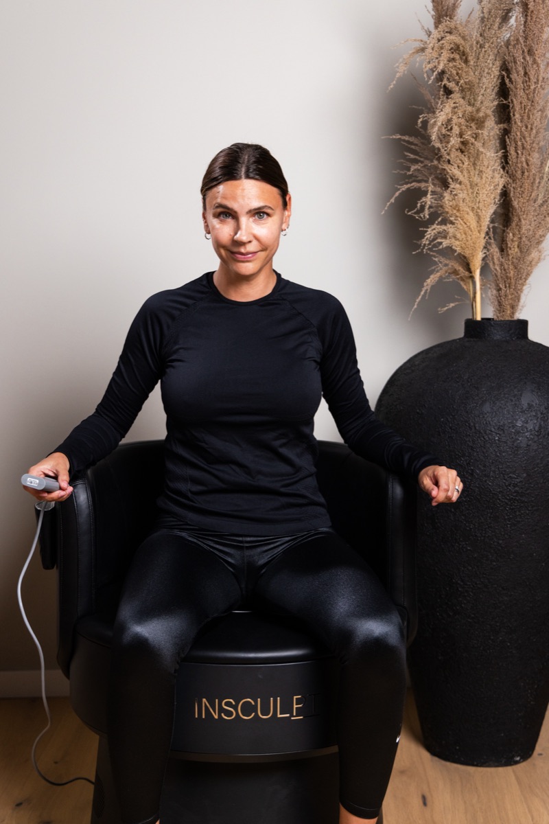 Kvinna sitter fullt påklädd i Insculpt Chair med handkontrollen i handen hos Studio Mione Clinic